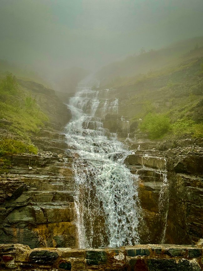 Tiered waterfall cascading down a rocky mountainside through thick fog in Glacier National Park