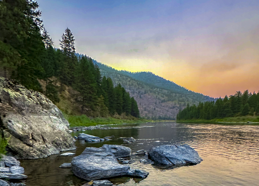 Large rocks resting in a calm Oregon river beneath evergreen-covered hills and a pastel sky