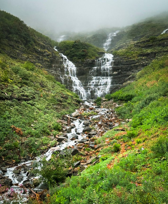 Two waterfalls cascading through green alpine vegetation and fog in Glacier National Park