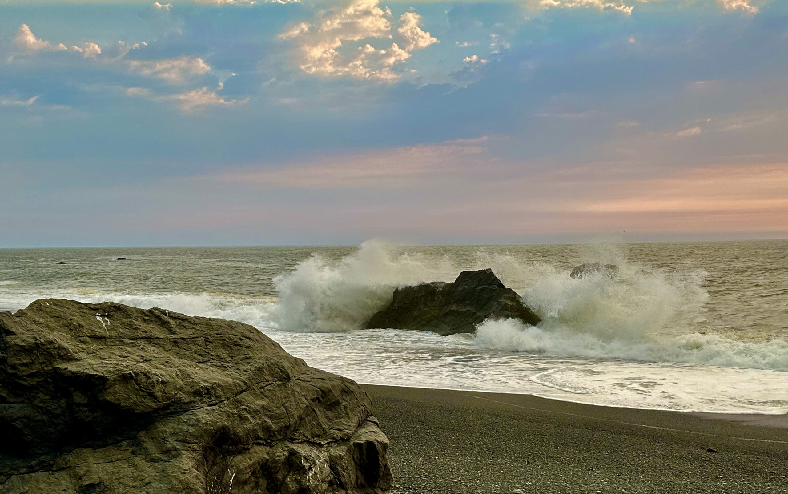 Pacific Ocean waves crashing against dark coastal rocks beneath pastel evening clouds near Klamath, California